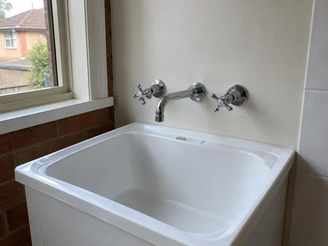 White laundry tub with chrome taps and spout beside a brick wall and window in West Ryde